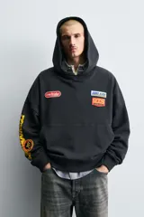 Sudadera tipo hoodie de corte relajado, color negro, con capucha y bolsillo frontal tipo canguro. Presenta parches decorativos en el pecho y estampados en color amarillo en la manga derecha. Acabados en rib en puños y ruedo.