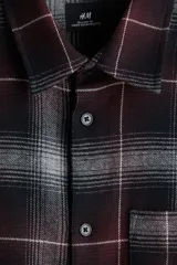 Camisa de franela de manga larga con cuello inglés, tapeta lisa, canesú en la espalda, bolsillo superior abierto y bajo redondeado. Presenta un patrón de cuadros en tonos negro, gris y granate.