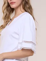 Remera blanca de silueta recta y algodón suave, con escote redondo y pequeña abertura central tipo gota. Presenta mangas de largo intermedio con aplicación horizontal de puntilla calada al tono y ruedo recto con tajos laterales.