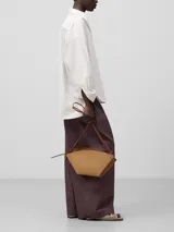 Bolso de hombro Adolfo Dominguez modelo Eclipse, confeccionado en piel bovina color tostado. Presenta un diseño estructurado formado por dos piezas asimétricas entrecruzadas, cierre con cremallera, asa de hombro extraíble y logo grabado en la parte frontal.