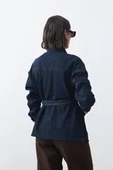 Sobrecamisa de jean color negro con silueta relajada, cuello camisero, cierre frontal con botones metálicos, bolsillos frontales y cinturón ajustable en la cintura.