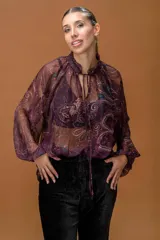 Blusa de gasa translúcida con estampado paisley en tonos burdeos, beige y azul. Presenta cuello alto con volante fruncido, abertura frontal con cordones ajustables y mangas largas abullonadas con puños elásticos.