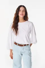 Blusa blanca de algodón, de corte recto y cuello redondo, con mangas largas abullonadas semitransparentes con textura calada de pequeñas flores y puños elásticos.