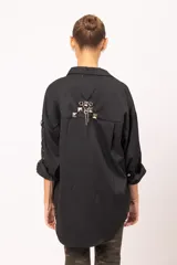 Camisa negra de microfibra con corte oversized. Presenta mangas con detalle de ojalillos y acordonado lateral, doble bolsillo frontal con solapa y botones metálicos. La parte trasera incluye diseño con tachas, ojalillos y cadena.