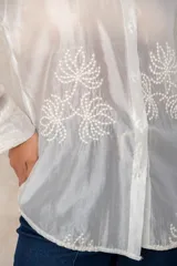 Camisa de manga larga en color blanco, confeccionada en tejido traslúcido con delicados bordados florales en hilo de seda al tono. Presenta cuello clásico, cierre frontal con botones y puños abotonados.