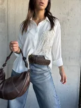 Camisa blanca de corte holgado con cuello clásico, cierre frontal de botones y detalle de bordado en relieve en el frente. Posee mangas largas con puños doblados.