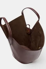 Bolso tipo shopper con forma de capazo, color marrón oscuro, confeccionado en material liso con pespuntes visibles en el cuerpo. Posee doble asa de mano y un asa bandolera ajustable. Incluye un bolso interior extraíble que se cierra mediante fruncido y un cierre principal con botón imantado.
