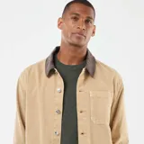 Chaqueta de trabajo (chore coat) color beige, confeccionada en tejido de algodón tipo sarga o drill, con cuello contrastante de pana marrón oscuro. Presenta cierre frontal de botones metálicos y cuatro bolsillos frontales de parche (dos superiores y dos inferiores).