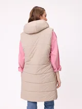 Chaleco largo acolchado color beige, con capucha forrada en sherpa, cierre frontal con cremallera y botones a presión, y bolsillos laterales.