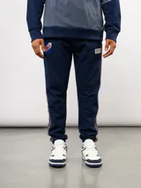 Pantalón deportivo de piqué color azul marino, con franjas laterales en contraste rojo y blanco. Presenta el escudo del Club Nacional de Football bordado en la pierna derecha y una etiqueta conmemorativa en la izquierda.