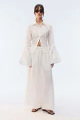 Camisa amplia de algodón liviano con textura calada, color blanco, cuello camisero, botones al frente, tiras para anudar en la espalda y mangas largas con puños acampanados y voluminosos.