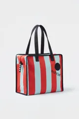 Tote bag de canvas con diseño a rayas verticales rojas y celestes, cierre superior y asas de hombro negras. Incluye la inscripción "PASTICHE" y un parche circular negro en el frente.