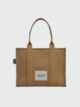 Bolso tipo tote de gran tamaño en color beige, confeccionado en lona resistente. Presenta el logo estampado en negro en el frente y detalles de cintas laterales con inscripciones. Cuenta con doble asa superior para llevar al hombro.