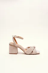 Sandalia de gamuza color beige con taco cuadrado, tira ajustable con hebilla en el tobillo y detalle de nudo en la tira delantera.