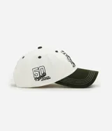 Gorra de béisbol de seis paneles, color blanco con visera curva en contraste de color verde militar. Presenta un bordado frontal de Snoopy junto a un banderín y el número cinco, y un bordado lateral con el texto 'Peanuts Athletic Department'.