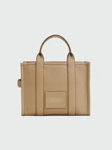 Cartera tipo tote de cuero granulado color tostado, con diseño estructurado y rectangular. Presenta doble asa superior fija y una correa larga ajustable y extraíble. En el frente, destaca el logo estampado con la inscripción 'THE TOTE BAG MARC JACOBS'.