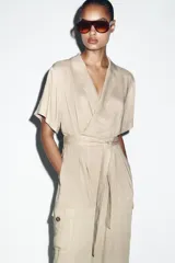 Mono largo de cuello solapa y escote en pico cruzado, con mangas cortas. Presenta bolsillos laterales de plastrón con solapa y botones, y un cinturón del mismo tejido para ajustar en la cintura. Cierre frontal con cremallera y botón interior.