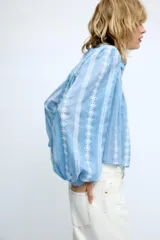 Blusa corta de color blanco con diseño de bordados perforados, cuello solapa, cierre frontal con botones y mangas largas abullonadas.