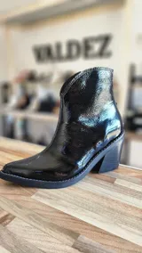 Botas tejanas de cuero charolado negro, con taco cuadrado de 7 cm y punta fina. Cuentan con cierre interno y costuras visibles en la suela.