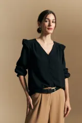 Camisa negra de manga larga con escote en V y cierre frontal de botones. Presenta detalles de volados en los hombros, frunces en la espalda y puños elastizados que le otorgan una silueta relajada y fluida.