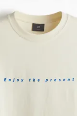 Remera de algodón color beige con cuello redondo y diseño holgado, con estampa frontal centrada con el texto "Enjoy the present" en azul.