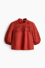 Blusa de viscosa color rojo óxido con secciones de broderie en el pecho y las mangas, cuello elevado festoneado y mangas globo al codo con puños elásticos.