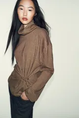 Top de cuello alto y manga larga, confeccionado en tejido de algodón gris melange. Presenta un diseño con frunces laterales ajustados mediante un aplique metálico decorativo.