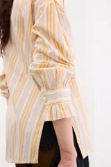 Camisa de corte holgado con estampado de rayas verticales en tonos amarillos, naranjas y beige. Presenta cuello alto con volados, cierre frontal con botones dorados texturados y mangas largas abullonadas con puños terminados en volados.