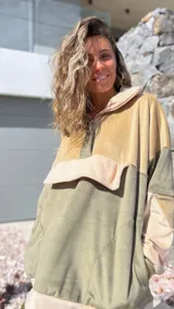 Buzo tipo canguro de diseño color block en tonos verde oliva y beige, confeccionado en tejido de textura aterciopelada. Presenta cuello alto con cierre frontal parcial, capucha y un bolsillo frontal grande con solapa.