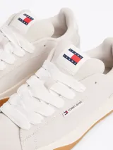Zapatillas Tommy Jeans de corte bajo, color gris claro, confeccionadas en ante con interior de poliéster y suela de goma color caramelo. Presentan cordones blancos, logo de la marca en la lengüeta y etiqueta de Tommy Jeans en el lateral.