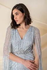 Vestido largo de tul en color celeste pastel, con escote en V y breteles anchos. Presenta un diseño bordado con lentejuelas y pedrería en líneas verticales y una capa larga integrada que nace desde los hombros.