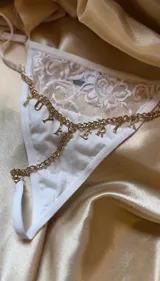 Tanga blanca con detalle de encaje floral en la parte superior y cadena dorada con dijes de letras colgantes.