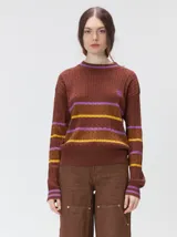 Sweater clásico de punto cable con cuello redondo a la base, mangas largas y fit al cuerpo. De color marrón con franjas horizontales violetas y amarillas, cuenta con el cuello, puños y ruedo acanalados, además de un pequeño logo bordado en el pecho.