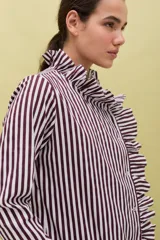 Camisa de algodón con estampado de rayas verticales en color bordó y blanco. Presenta un diseño de manga larga con puños abotonados, cuello camisero y un detalle de volado lateral que recorre el frente desde el cuello hasta el ruedo, acompañando la línea de botones.