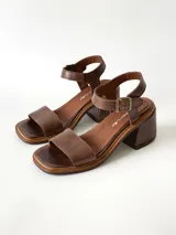 Sandalias de cuero color chocolate, con taco cuadrado de 6 cm, tira ancha en la parte delantera y tira en el tobillo con hebilla metálica ajustable.