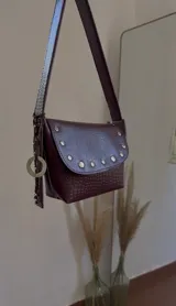 Cartera bandolera color bordó con textura símil cocodrilo, solapa con tachas doradas y correa larga regulable. Incluye charm circular plateado.