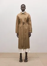 Chaqueta larga de algodón color marrón tostado, estilo gabardina, con cuello camisero, cierre frontal oculto con botones, mangas largas con presillas en los puños y cinturón ajustable con hebilla oscura.