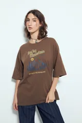 Remera blanca de manga corta con calce relajado y cuello a la base. Presenta un estampado frontal pequeño y una estampa protagonista en la espalda con el texto "Rocky Mountain National Park" y un paisaje de montañas y árboles.