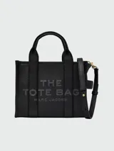 Cartera tipo tote de cuero rojo de la marca Marc Jacobs, modelo The Leather Small Tote Bag. Presenta doble asa superior, correa ajustable y extraíble para el hombro, y el texto grabado en relieve "THE TOTE BAG MARC JACOBS" en el frente.