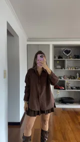 Camisa de corte oversize en color marrón con estampado de rayas verticales finas en tono claro. Presenta cuello clásico, cierre frontal con botones y bolsillo en el pecho.