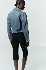 Cazadora de denim estilo crop con cuello solapa, manga larga y cierre frontal con cremallera y botón metálico. Presenta bolsillos de vivo en el delantero y detalle de trabillas en los hombros.