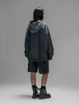 Hoodie oversize de color gris oscuro, confeccionado en frisa de algodón con recortes en textiles técnicos. Presenta capucha forrada, bolsillo frontal tipo canguro y un bolsillo adicional con solapa en la parte inferior.