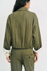Chaqueta corta de color verde militar, con cuello solapa, bolsillos delanteros con solapa y cierre frontal de botones ocultos. Presenta mangas amplias con vuelta en los puños que muestran un forro a rayas.
