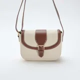 Bolso bandolera de diseño estructurado, confeccionado en lona color beige con detalles de cuero sintético marrón en los bordes y la solapa frontal. Cuenta con una hebilla decorativa metálica, costuras a la vista en color blanco y correa ajustable.