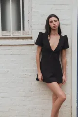 Vestido mini negro con escote en V amplio, mangas cortas abullonadas y detalle de lazo en el delantero. Cuenta con cierre de cremallera oculta en la espalda.
