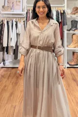 Vestido largo de seda en color beige, con mangas tres cuartos, cuello camisero y falda amplia con caída fluida. Incluye un cinturón tejido en tono marrón que marca la cintura.