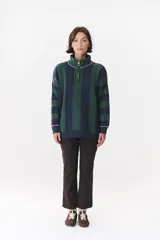 Buzo de punto con diseño de rayas verticales en tonos verde y azul marino. Presenta cuello alto con cierre frontal, hombros caídos y coderas en contraste de color marrón con costuras decorativas visibles.