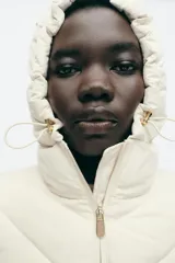 Cazadora de mujer con diseño híbrido, presenta un panel frontal acolchado con costuras en chevron y mangas y laterales en tejido liso. Cuenta con cuello alto, capucha ajustable con cordones elásticos y cierre frontal mediante cremallera.