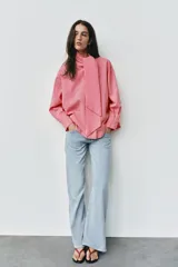 Camisa de rayas rosadas y blancas, con cuello subido decorado con un lazo integrado, manga larga con puños abotonados y cierre frontal oculto.