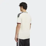 Camiseta de fútbol Adidas del Manchester United, correspondiente a la tercera equipación. Es de color blanco marfil con detalles en bordó y negro en el cuello, puños y hombros. Cuenta con el logotipo de Adidas, el escudo del club y un gráfico alineados verticalmente en el centro del pecho.